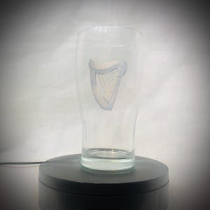 Puede incluir: Un vaso de pinta transparente con un logotipo de arpa azul claro y blanco. El vaso tiene forma cil&iacute;ndrica con una ligera abertura en la parte superior. Est&aacute; sobre una base giratoria negra.