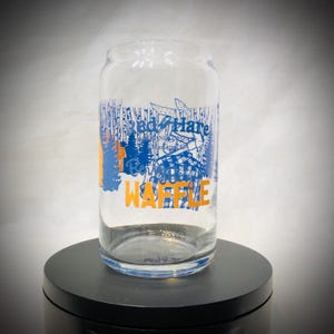 Könnte beinhalten: Ein klares, glasförmiges Trinkglas mit blauen und orangefarbenen Grafiken. Der Text auf dem Glas lautet "Red Hare Brewing" und "Waffle". Das Design beinhaltet eine Waldszene. Das Glas steht auf einer schwarzen Oberfläche.