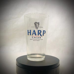 Puede incluir: Un vaso de pinta transparente con el logotipo de Harp Lager. El logotipo presenta un arpa sobre la palabra "HARP" en letras azules y doradas. El texto "LAGER" est&aacute; debajo de la palabra "HARP". El vaso est&aacute; sobre una superficie negra.