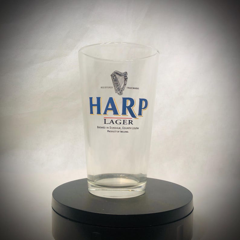 Puede incluir: Un vaso de pinta transparente con el logotipo de Harp Lager. El logotipo presenta un arpa sobre la palabra "HARP" en letras azules y doradas. El texto "LAGER" est&aacute; debajo de la palabra "HARP". El vaso est&aacute; sobre una superficie negra.