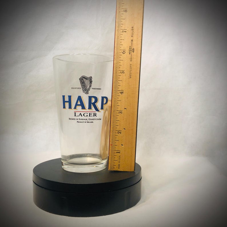 Puede incluir: Un vaso de pinta transparente de Harp Lager con el logotipo y el texto de Harp en azul y blanco. Una regla de madera se coloca junto al vaso. El vaso est&aacute; sobre un soporte giratorio negro. El vaso mide unos 15 cm de alto.