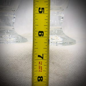 May include: Two clear glass pedestal goblets positioned behind a yellow measuring tape. The tape shows the glass base and stem structure, with the visible portion of the tape spanning from the 5-inch mark to the 8-inch mark on the scale.