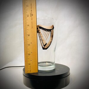 Puede incluir: Un vaso de pinta transparente con un logotipo de arpa, de aproximadamente 15 cm de altura, se encuentra sobre una base giratoria negra. Una regla de madera est&aacute; colocada junto al vaso. El logotipo del arpa es marr&oacute;n y negro.