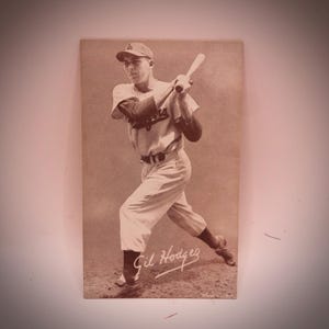 Könnte beinhalten: Sepiafarbene Vintage-Baseballkarte mit Gil Hodges in Schlaghaltung. Er trägt eine Dodgers-Uniform, eine Kappe und hält einen Schläger. Die Karte hat das Dodgers-Logo und die Unterschrift von Gil Hodges.