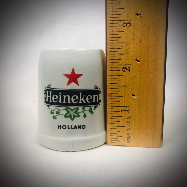 May include: A white ceramic shot glass with the Heineken logo, a red star, and the word "Holland". The shot glass is approximately 2 inches tall. A wooden ruler is next to the shot glass.
