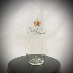 May include: A clear, tall champagne flute with the words "Grand Duval" in elegant script. The glass features a crest design in gold, red, and white. The glass is displayed on a black surface.