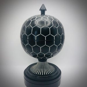 Puede incluir: Una esfera decorativa negra con un patrón geométrico de panal y un pequeño remate en forma de flecha. La esfera se asienta sobre una base de pedestal con un diseño radial. El diseño general es simétrico y elegante.