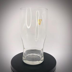 May include: A clear pint glass with a slightly flared shape. A gold harp logo is visible on the front of the glass. The glass is empty and sits on a black surface.