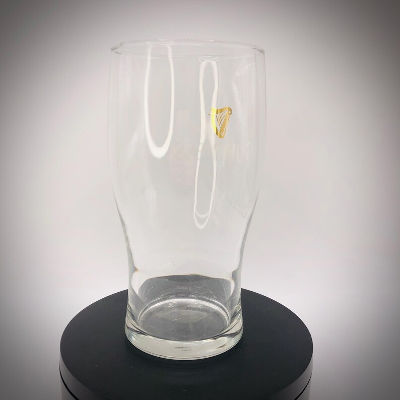 May include: A clear pint glass with a slightly flared shape. A gold harp logo is visible on the front of the glass. The glass is empty and sits on a black surface.