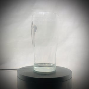 Puede incluir: Un vaso de cerveza claro y alto, con forma curva y base gruesa. El vaso tiene un dise&ntilde;o sutil en el lateral y est&aacute; vac&iacute;o. Se encuentra sobre una plataforma giratoria negra sobre un fondo blanco.