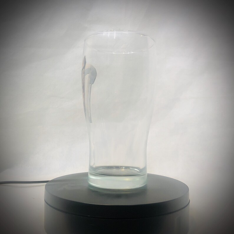 Puede incluir: Un vaso de cerveza claro y alto, con forma curva y base gruesa. El vaso tiene un dise&ntilde;o sutil en el lateral y est&aacute; vac&iacute;o. Se encuentra sobre una plataforma giratoria negra sobre un fondo blanco.