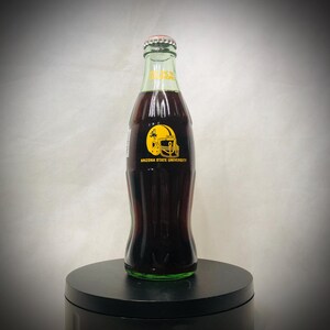 Könnte beinhalten: Eine Glasflasche dunkler Limonade mit rotem und silbernem Verschluss. Die Flasche hat ein gelbes Logo eines Footballhelms der Arizona State University. Die Flasche steht auf einem schwarzen Ständer.