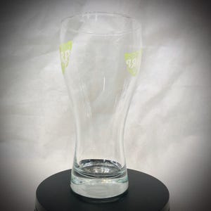 May include: A clear, tulip-shaped beer glass with a wide base and a slightly flared top. The glass has a green logo on both sides. The glass is empty and sits on a black surface.