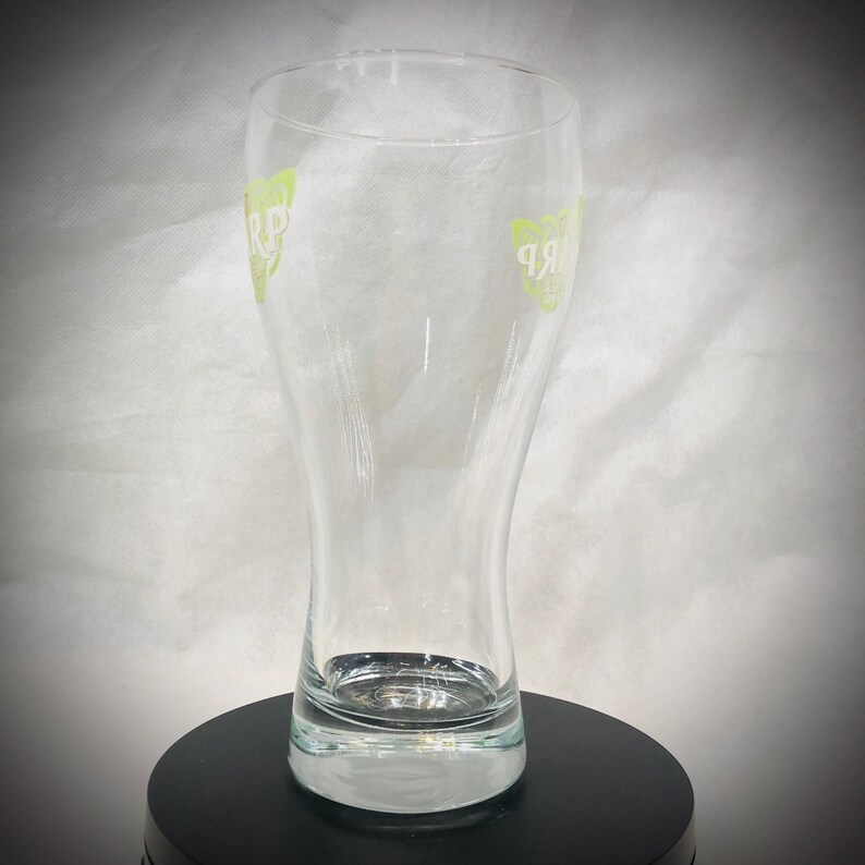 May include: A clear, tulip-shaped beer glass with a wide base and a slightly flared top. The glass has a green logo on both sides. The glass is empty and sits on a black surface.