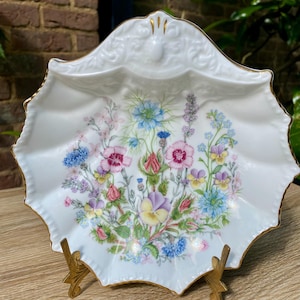 May include: A white porcelain dish with a scalloped edge and gold trim. The dish is decorated with a floral design in pink, blue, purple, and yellow. The dish is displayed on a gold-coloured metal stand.