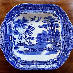 May include: Blue and white ceramic serving dish with a Willow pattern design. The dish has a square shape with rounded corners and a decorative border. The central image depicts a landscape with a bridge, trees, and buildings.