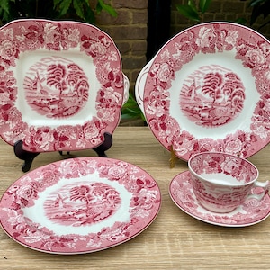 May include: A vintage pink and white porcelain tea set with a landscape design. The set includes a square serving plate, a round serving plate, a small oval plate, a teacup, and a saucer. The plates and cup have floral borders.