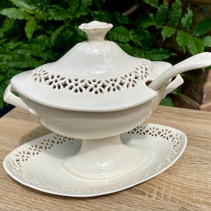 May include: A cream-coloured ceramic soup tureen with a matching lid and spoon. The tureen has a pedestal base and an oval serving plate. The lid features a decorative floral pattern and a small handle. The spoon rests on the side.