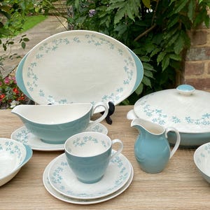 May include: A collection of vintage ceramic dinnerware in white and teal. The set comprises a serving platter, a lidded dish, a gravy boat, a creamer jug, a cup and saucer, and two bowls. Each item is decorated with a floral pattern.