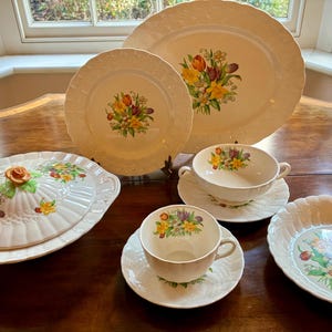 May include: A vintage cream porcelain tableware set featuring a floral design. The set includes plates, bowls, a covered dish, and teacups, arranged on a wooden table. The floral pattern features yellow, red, and purple flowers.