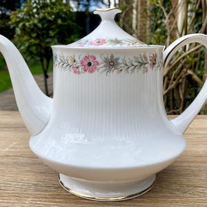 May include: White porcelain teapot with a lid, spout, and handle. The teapot has a vertical ribbed design and a floral band with pink and blue flowers. Gold trim accents the base and lid.