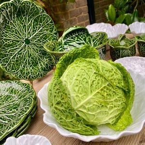 May include: Ceramic tableware set featuring a cabbage leaf design. The collection includes green and white plates, bowls, and lidded dishes. A fresh cabbage is presented in a white, scalloped bowl. Measurements are in cm.