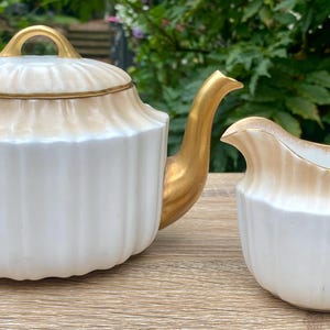 May include: A white porcelain teapot and creamer set with gold detailing. The teapot has a lid and a curved spout, and the creamer has a handle. Both items feature a ribbed design and a tan band at the top.