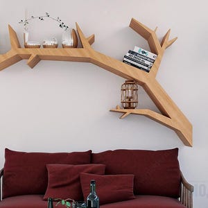 May include: A wooden tree-shaped shelf is mounted on a white wall, holding decorative items and books. Below, a burgundy sofa with matching pillows sits against the wall. A decorative lantern and a potted plant complete the scene.