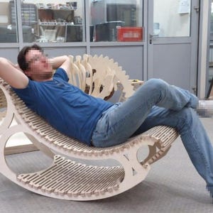 Op de afbeelding: Een lichtgekleurde houten schommelstoel met een uniek, gebogen ontwerp. De stoel heeft een lattenzitting en rugleuning, en een decoratief, open ontwerp op de rug. De stoel is ontworpen voor comfort en ontspanning.