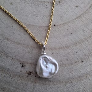 Könnte beinhalten: Eine goldene Halskette mit einem großen, unregelmäßig geformten weißen Perlenanhänger. Die Perle hat eine einzigartige, organische Form und eine glänzende Oberfläche. Die Halskette wird auf einem hellbraunen Holzgrund präsentiert.