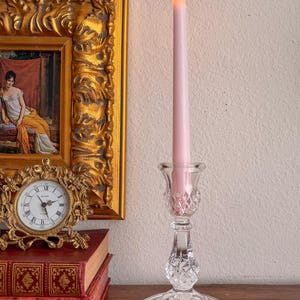 Könnte beinhalten: Eine brennende, hellrosa Kerze in einem durchsichtigen Glas-Kerzenhalter. Der Halter hat ein Rautenmuster am Stiel und einen runden Fuß. Ein goldgerahmtes Gemälde und eine kleine Uhr sind im Hintergrund.