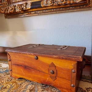 Può includere: Un piccolo forziere antico in legno con coperchio incernierato e ferramenta in metallo. Il forziere è realizzato in legno marrone chiaro con accenti più scuri e borchie decorative in metallo. È appoggiato su una superficie a motivi.