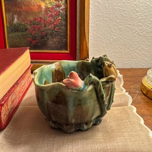Może przedstawiać: Ceramiczna miska z motywem żaby i lilii wodnej. Misa jest głównie zielona z brązowymi i niebieskimi akcentami. Różowy kwiat znajduje się z boku. Misa stoi na beżowej powierzchni.