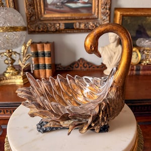 Puede incluir: Un cuenco decorativo con forma de cisne que presenta un cuerpo de cristal transparente con detalles de plumas moldeadas y un cuello y base en tono dorado metálico. La cabeza del cisne está curvada hacia abajo y las alas forman los lados.