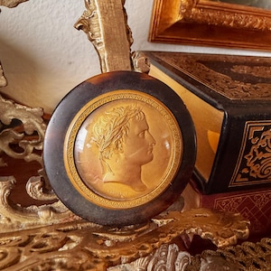 May include: A round, antique-style decorative object with a portrait of a man in profile, wearing a laurel wreath. The portrait is set within a dark brown, circular frame. The background includes a gold-framed painting and a decorative gold stand.