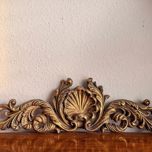 May include: A horizontal gold-colored wall ornament featuring a central scallop shell motif flanked by symmetrical, intricate acanthus leaf scrollwork. The piece has a textured, antiqued finish with deep shadows highlighting the relief details.