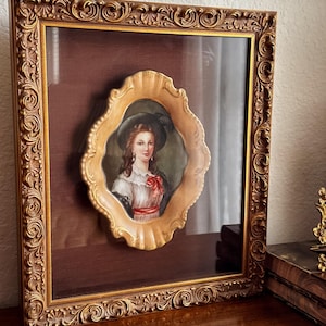 May include: Ornate gold-colored framed portrait of a woman in a hat. The portrait is oval-shaped with a detailed gold frame, set against a dark red background. The outer frame is also gold, with intricate scrollwork.