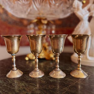 Puede incluir: Cuatro copas antiguas de latón con bordes acampanados y tallos ornamentados se colocan sobre una superficie de mármol verde oscuro. Las copas tienen un tono dorado cálido, que refleja la luz. El fondo incluye elementos decorativos borrosos.