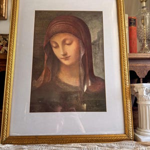 May include: A framed portrait of a woman with a red head covering, her eyes closed, in a gold ornate frame. The artwork has a soft, muted color palette with a focus on the woman's face. The frame is rectangular and has a detailed, textured design.