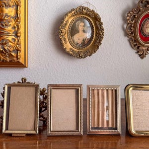 May include: A collection of eight small picture frames arranged in a row, featuring various metallic finishes including gold, brass, and silver tones. The frames vary in shape, including rectangular and oval designs, with some containing textured inserts.