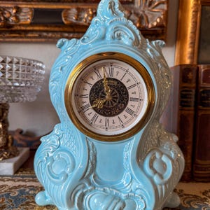 May include: A light blue ceramic mantel clock with a gold-tone clock face. The clock has ornate detailing and Roman numerals. The clock face has the word "Marco" printed on it.