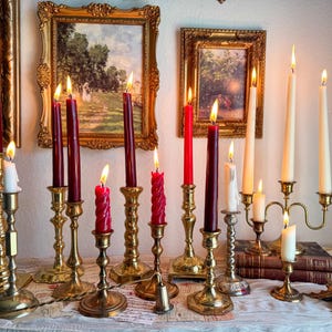 May include: An array of lit candles in shades of red, burgundy, and white, presented in decorative brass and gold candle holders. The candles are arranged on a table, with antique gold framed artwork in the background.