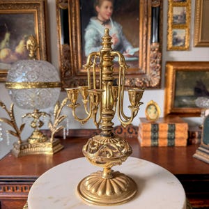 May include: A gold-toned metal candelabra featuring a central pillar, a decorative openwork base, and four curved arms extending outward to hold candles. The structure is finished with a fluted circular base and a pointed finial at the top.