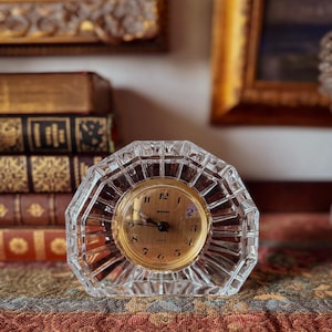 May include: A clear, faceted crystal quartz clock with a gold-tone face. The clock has a geometric, octagonal shape and displays the time with black hands and gold numerals. The clock is set against a backdrop of antique books and framed artwork.