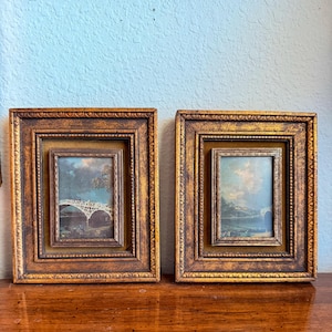 May include: Two rectangular wall frames with ornate, distressed gold-toned borders and dark brown inner matting. Each frame contains a small landscape print featuring a white arched bridge over water under a cloudy sky, set within a recessed frame.