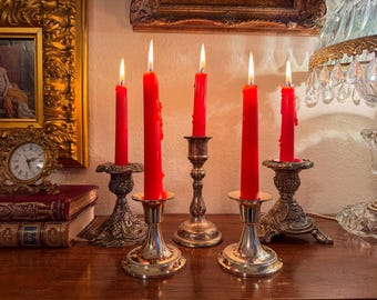 Può includere: Cinque candele rosse accese, con cera che cola, poste in candelabri argentati e bronzati ornati. Le candele sono su una superficie di legno, con un quadro incorniciato e un orologio sullo sfondo. La scena evoca un'estetica vintage.