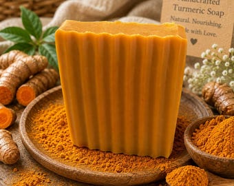 May include: A rectangular bar of orange turmeric soap with a vertically ribbed texture, resting on a wooden plate covered in turmeric powder, surrounded by raw turmeric roots and a card labeled Handcrafted Turmeric Soap Natural Nourishing.