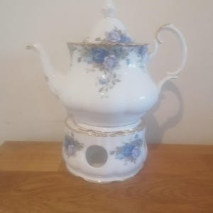 Peut inclure: Théière en porcelaine blanche avec chauffe-plat assorti. La théière présente un motif floral avec des roses bleues et des bordures dorées. Le chauffe-plat, avec une ouverture circulaire, reprend le motif floral. La théière a un couvercle et une anse incurvée.