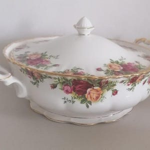 May include: A white ceramic serving dish with a lid, decorated with a rose floral pattern in red, pink, and yellow. The dish has gold trim and handles, and a decorative finial on the lid. A classic piece for serving.