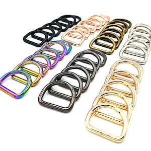 Puede incluir: Surtido de anillas en D en varios colores, incluyendo negro, oro rosa, bronce, plata, oro e iridiscente. Estas anillas en D metálicas están dispuestas en filas, mostrando sus diferentes acabados y estilos. Ideal para manualidades y fabricación de bolsos.